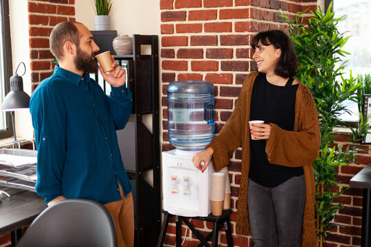 Businessman taking work break talking with executive manager collaborating and communicating in startup office drinking cup of water as refreshment. Businesspeople sitting near to water cooler