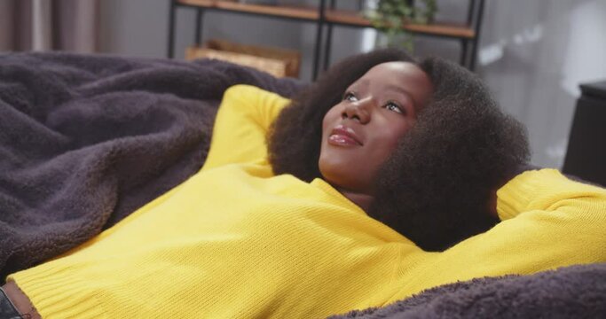 Side shot of african american wooman lying on sofa with arms crossed behing her head looking onto ceiling. Female in yellow clothes. Tired female after work resting. Concept of rest, carefree.