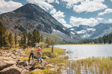 Two girls travelers walks along a trail in a national park with a mountain river and lake in the background.