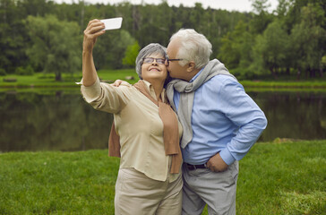 Cheerful old wife making selfie by phone camera while tender loving husband sweet kissing her cheek. Retired people walking in city public park. Good memory and best vibes on pension concept