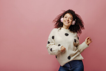Fototapeta na wymiar Smiling young caucasian lady is enjoying her favorite song and dancing on pink background. Portrait of curly brunette woman having fun in headphones. Audio technology, and music concept.