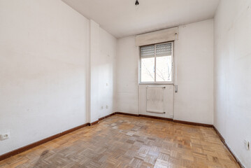 empty living room with white aluminum radiator under a window and dirty oak parquet flooring