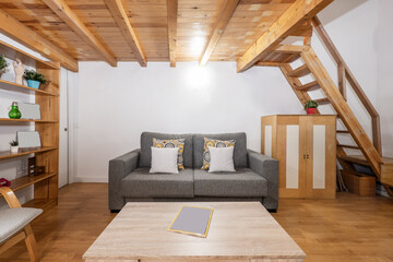 Living room with light wood coffee table, sofa bed upholstered in gray fabric, shelves and wooden staircase and wooden floors
