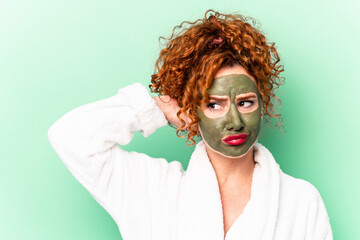 Young red hair woman with a facial aloe vera mask after a bath isolated touching back of head, thinking and making a choice.