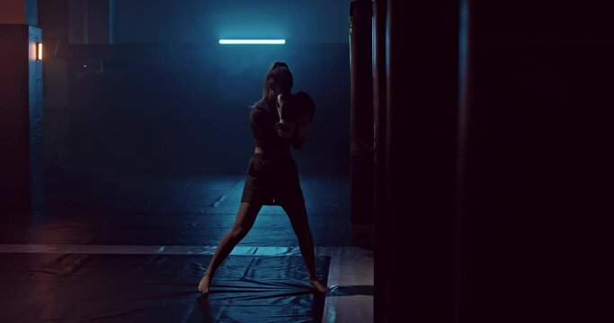 Female athlete boxing a punching bag in a dark hall of a fight club, punching bags hang in a row. Athlete professionally trains before the fight, practicing punches on a punching bag, silhouette. 4k