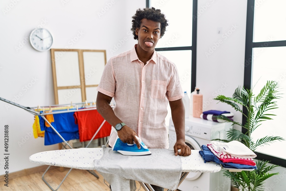 Poster african man with curly hair ironing clothes at home sticking tongue out happy with funny expression.