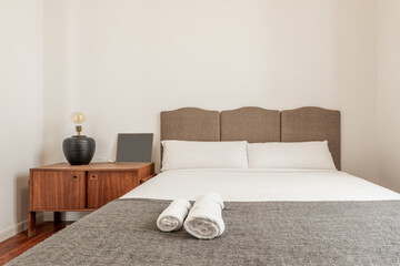 Bedroom with a king-size bed with a fabric-upholstered headboard, a wooden bedside table and a lamp without a shade and rolled-up white towels