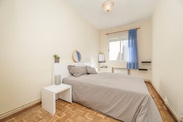 Bedroom with king size bed with gray bedspread, round mirror and aluminum window with blue curtain