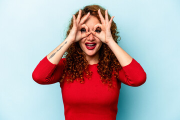 Young ginger caucasian woman isolated on blue background showing okay sign over eyes