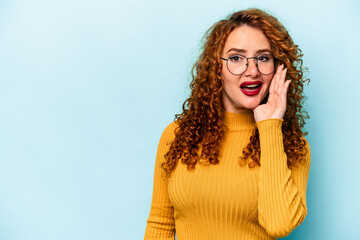Young ginger caucasian woman isolated on blue background shouts loud, keeps eyes opened and hands tense.