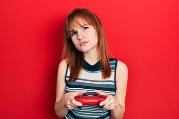 Redhead young woman playing video game holding controller clueless and confused expression. doubt...