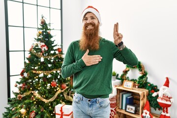 Redhead man with long beard wearing christmas hat by christmas tree smiling swearing with hand on...