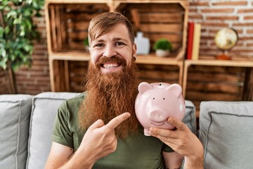 Redhead man holding piggy bank smiling happy pointing with hand and finger