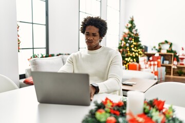 Young african american man using laptop sitting on the table by christmas tree puffing cheeks with...