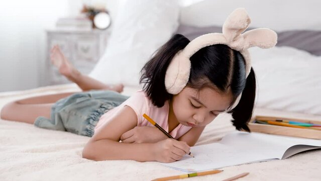 4K 50fps, adorable Asian girl tied her hair and wearing bunny ears, happily laying on a white bed drawing pictures.