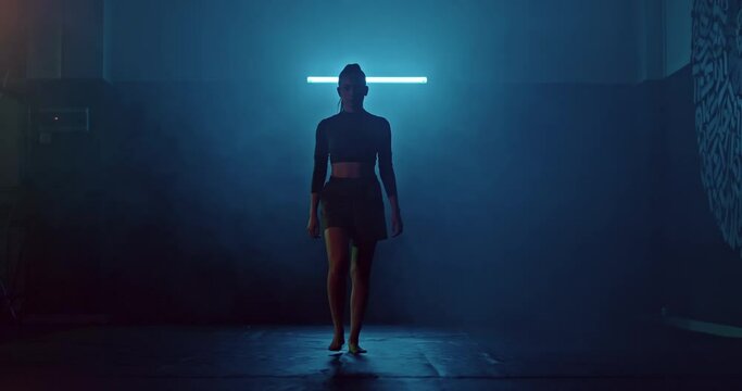 Silhouette, a young female athlete walks through the dark hall of the fight club, tubers of smoke. Woman is preparing for a fight, a serious look. Modern fighting hall, punching bags hanging in a row