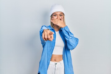 Beautiful blonde woman wearing wool hat laughing at you, pointing finger to the camera with hand over mouth, shame expression