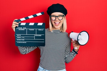 Beautiful blonde woman holding video film clapboard and megaphone smiling and laughing hard out...