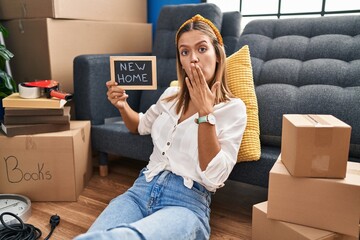 Young blonde woman holding new home blackboard covering mouth with hand, shocked and afraid for...