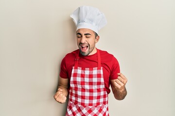 Young hispanic man wearing baker uniform excited for success with arms raised and eyes closed...