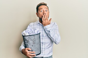 Middle age man holding paper bin full of crumpled papers covering mouth with hand, shocked and...