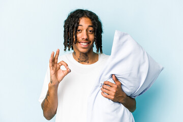 Young African American man holding a pillow isolated on blue background cheerful and confident...
