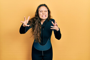 Young hispanic girl wearing diver neoprene uniform shouting frustrated with rage, hands trying to...