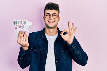 Young hispanic man holding egyptian pounds banknotes doing ok sign with fingers, smiling friendly gesturing excellent symbol - obrazy, fototapety, plakaty