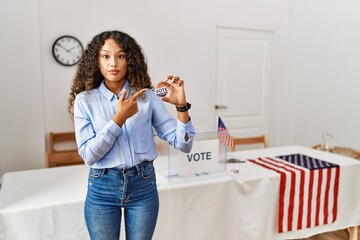 Beautiful hispanic woman standing by at political campaign by voting ballot pointing with hand...