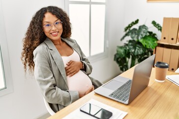 Young latin woman pregnant smiling confident touching tummy working at office