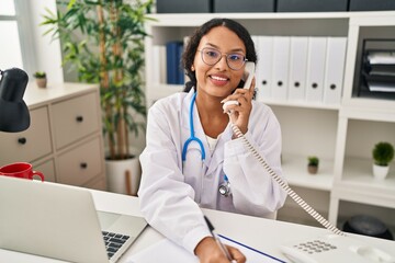 Young latin woman wearing doctor uniform talking on the telephone write on clipboard at clinic