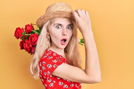 Beautiful caucasian woman with blond hair holding bouquet of red roses afraid and shocked with surprise and amazed expression, fear and excited face.