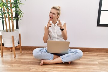 Young blonde woman using computer laptop sitting on the floor at the living room pointing fingers to camera with happy and funny face. good energy and vibes.