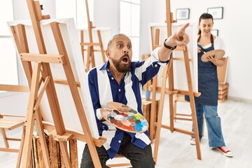Senior artist man at art studio pointing with finger surprised ahead, open mouth amazed expression, something on the front