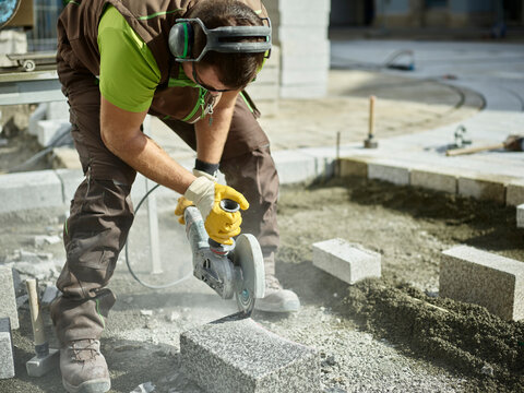 Blue-collar worker cutting paving stone by electric saw at construction site