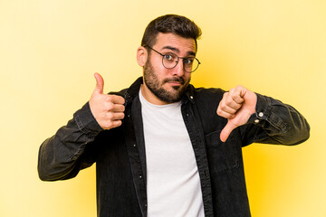 Young caucasian man isolated on yellow background showing thumbs up and thumbs down, difficult...