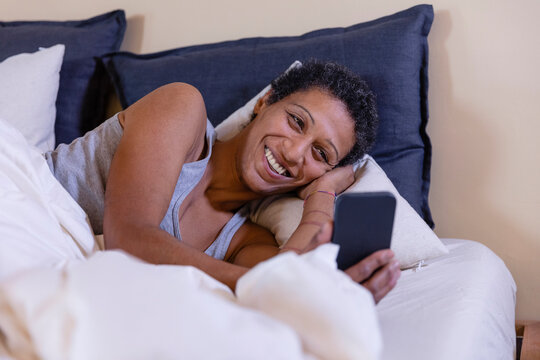 Smiling woman using mobile phone lying on bed in bedroom