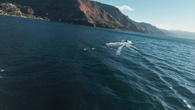 Boat speeding through waves of lake Atilan on sunny day. Mountain range in background. Aerial fpv view
