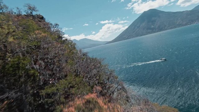 Fly close to bushy cliff side revealing majestic lake Atilan with boat and volcano in background, aerial FPV view