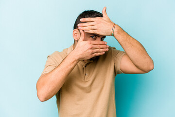 Fototapeta premium Young caucasian man isolated on blue background blink at the camera through fingers, embarrassed covering face.