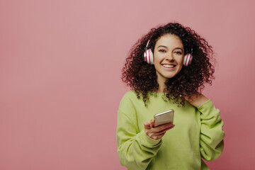 Beautiful young fair-skinned girl in headphones holds smartphone, selects track in streaming application, standing on pink background. Curly brunette in casual jacket listens to music. 