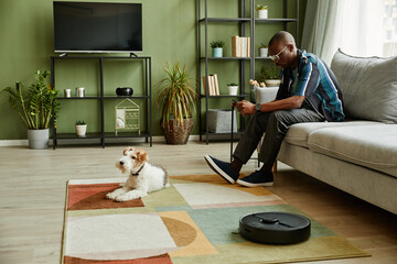 Side view portrait of modern African American man relaxing on couch at home with dog and robot...