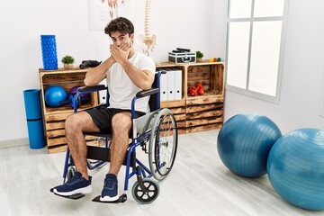 Fototapeta premium Hispanic man sitting on wheelchair at physiotherapy clinic shocked covering mouth with hands for mistake. secret concept.