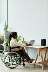 Minimal full length of young black woman in wheelchair using laptop while working at home office,...