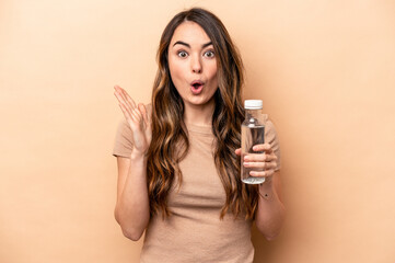 Young caucasian woman holding a bottle of water isolated on beige background surprised and shocked.