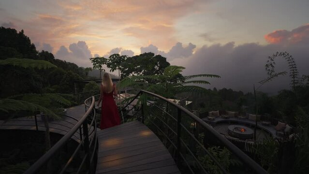 Woman in red dress walks wooden boardwalk above tree canopy during magical sunset