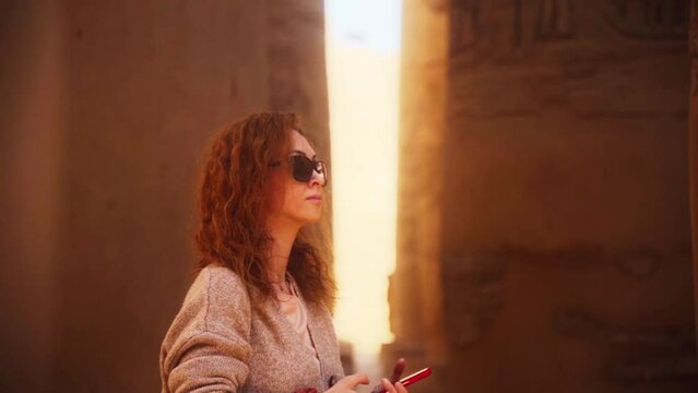 Female tourist taking a picture with her phone in the Karnak temple, Luxor Egypt. 
Shallow depth of field, slow movement, warm colours and the bright sun backlighting the woman between two pillars.