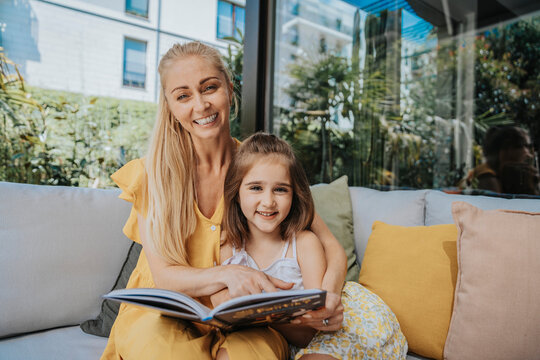 Smiling mother and daughter with book on sofa at patio