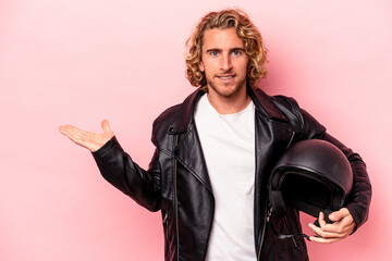 Young caucasian man with a motorcycle helmet isolated on pink background showing a copy space on a palm and holding another hand on waist.