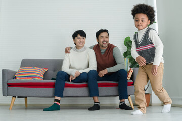 Fototapeta premium Happy African adopted kid boy wearing sweater dancing Hip Hop in front of Asian young gay parent looking with pride face at living room at home. Lgbt family supporting dream of their son